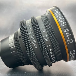 Puede incluir: Un objetivo Helios 44-2 de 58 mm negro con un anillo dorado alrededor del barril. El objetivo tiene una escala de profundidad de campo marcada con números del 2,8 al 16.