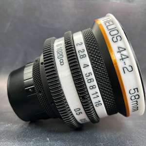 Op de afbeelding: Een zwarte camera lens met witte markeringen. De lens heeft een gouden band eromheen en de tekst "HELIOS 44-2 58mm" staat op de zijkant.