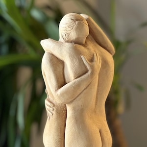 Può includere: Scultura beige raffigurante due figure che si abbracciano. Le figure hanno forme lisce e arrotondate, con le braccia avvolte l'una all'altra. Lo sfondo è sfocato, con una pianta visibile.