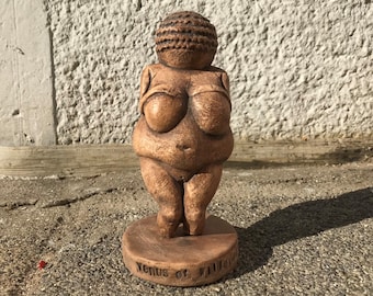 Peut inclure: Une sculpture en céramique brune de la Vénus de Willendorf, une figurine préhistorique représentant une femme aux traits exagérés. La sculpture est sur un socle avec les mots "Vénus de Willendorf" inscrits dessus.