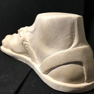K&ouml;nnte beinhalten: Eine wei&szlig;e Gips-Skulptur eines Fu&szlig;es, der eine Sandale tr&auml;gt. Die Sandale hat einen Riemen, der um den Kn&ouml;chel geht, und eine dicke Sohle.