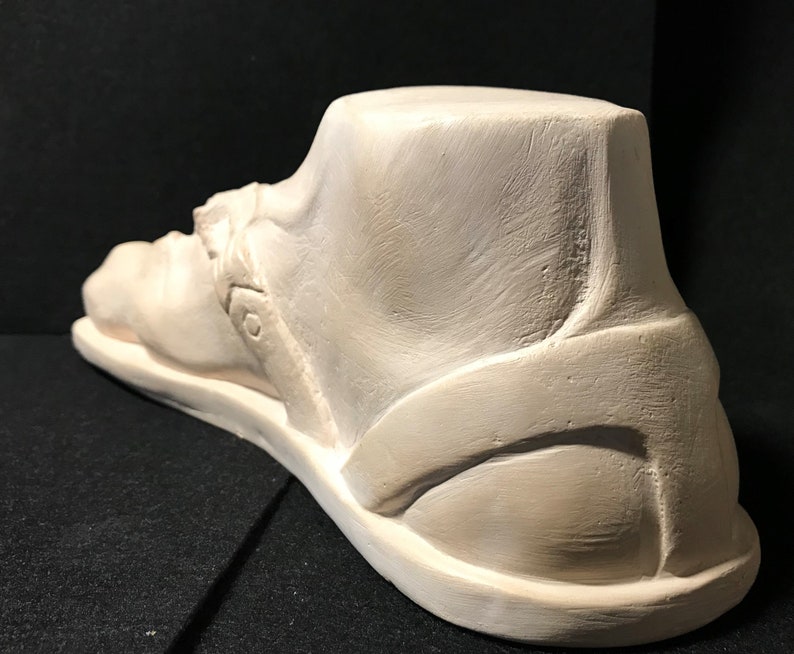 K&ouml;nnte beinhalten: Eine wei&szlig;e Gips-Skulptur eines Fu&szlig;es, der eine Sandale tr&auml;gt. Die Sandale hat einen Riemen, der um den Kn&ouml;chel geht, und eine dicke Sohle.