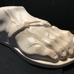 K&ouml;nnte beinhalten: Eine wei&szlig;e Gips-Skulptur eines Fu&szlig;es, der eine Sandale mit einem detaillierten Design tr&auml;gt. Die Sandale hat einen Riemen, der um den Kn&ouml;chel geht, und eine dekorative Schnalle.