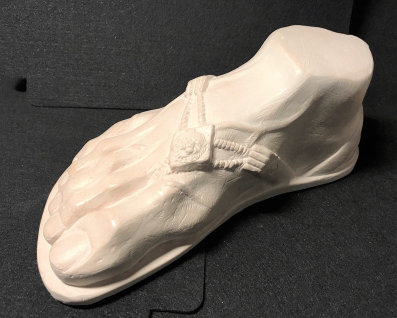 K&ouml;nnte beinhalten: Eine wei&szlig;e Gips-Skulptur eines Fu&szlig;es, der eine Sandale tr&auml;gt. Die Sandale hat einen Riemen mit einer dekorativen Schnalle.