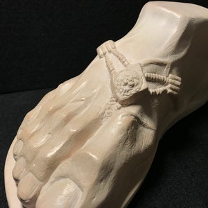 K&ouml;nnte beinhalten: Eine wei&szlig;e Gips-Skulptur eines Fu&szlig;es mit einem detaillierten Design einer Kette und einem L&ouml;wenkopf am Kn&ouml;chel.