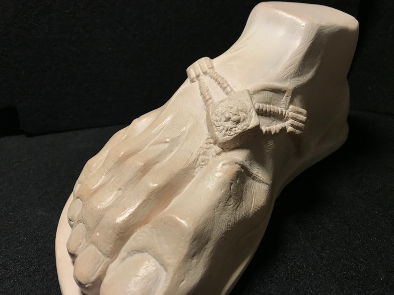 K&ouml;nnte beinhalten: Eine wei&szlig;e Gips-Skulptur eines Fu&szlig;es mit einem detaillierten Design einer Kette und einem L&ouml;wenkopf am Kn&ouml;chel.