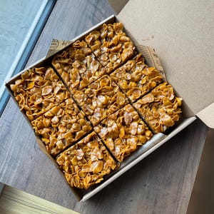 May include: A box of 12 golden brown squares of cereal bars with a caramel-like topping. The bars are arranged in a grid pattern.