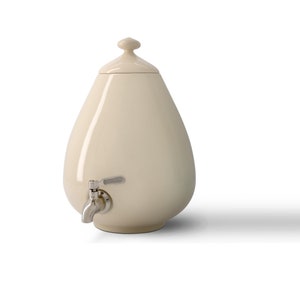 May include: Cream-colored ceramic beverage dispenser with a lid and a silver-toned spigot. The dispenser has a rounded, teardrop shape and sits on a white surface. The lid has a small, rounded handle.