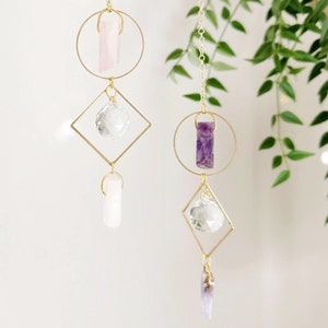 May include: Two hanging crystal suncatchers with gold-colored metal frames. Each suncatcher features a combination of crystals in various shapes and colors, including rose quartz, amethyst, and clear quartz. The design includes circles, squares, and dangling crystal pendants.