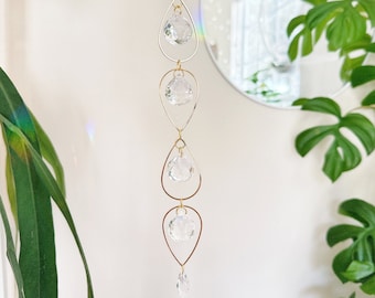 May include: A gold crystal suncatcher with five clear crystal drops hanging from a chain. The suncatcher is hanging from a ceiling in front of a mirror.