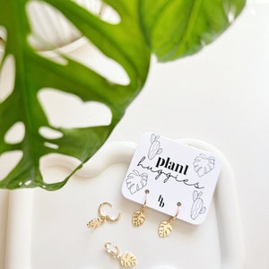 May include: Three pairs of gold-toned earrings with a leaf design. The earrings are displayed on a white card with the text "plant huggies".