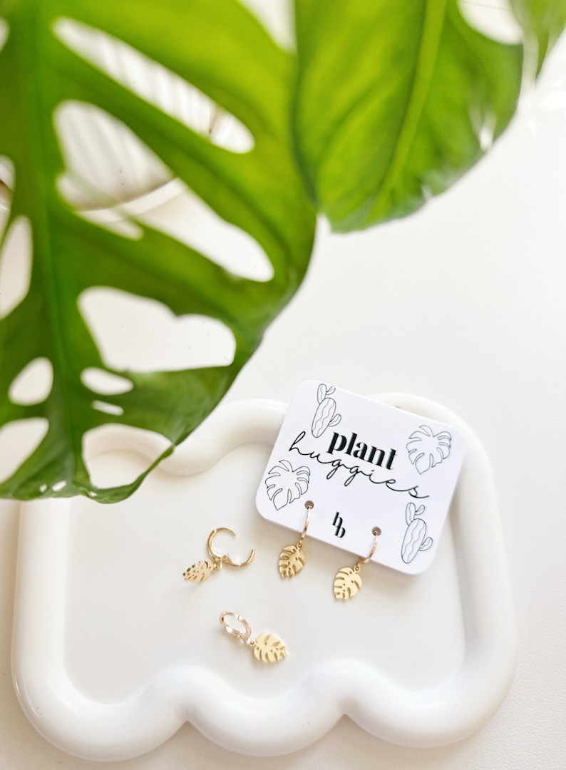 May include: Three pairs of gold-toned earrings with a leaf design. The earrings are displayed on a white card with the text "plant huggies".
