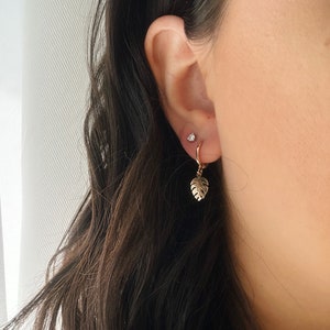 May include: Gold hoop earrings with a small, delicate leaf charm hanging from one earring. The earrings are worn in a person's ear.