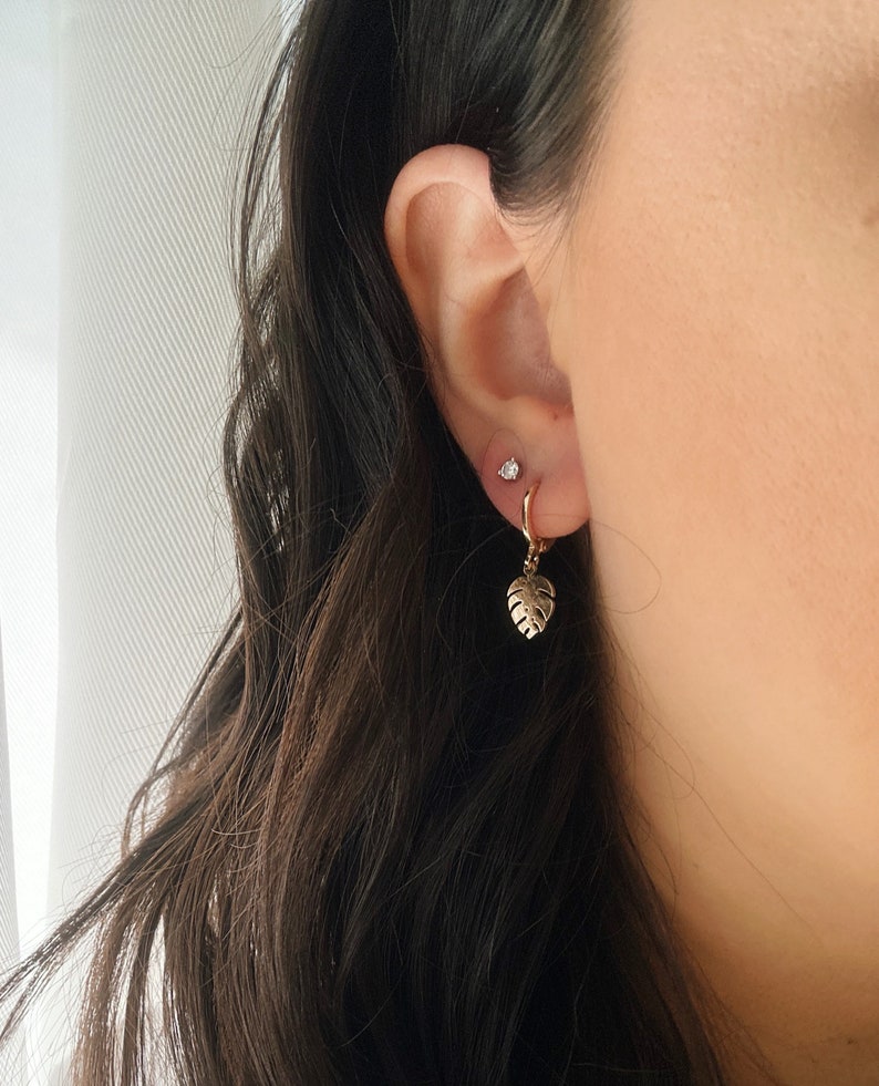 May include: Gold hoop earrings with a small, delicate leaf charm hanging from one earring. The earrings are worn in a person's ear.
