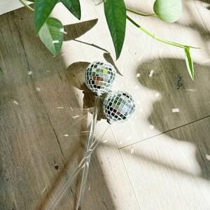May include: Two silver disco ball ornaments on clear acrylic sticks. The sticks are crossed and the ornaments are facing each other. The ornaments are on a light brown wood floor.