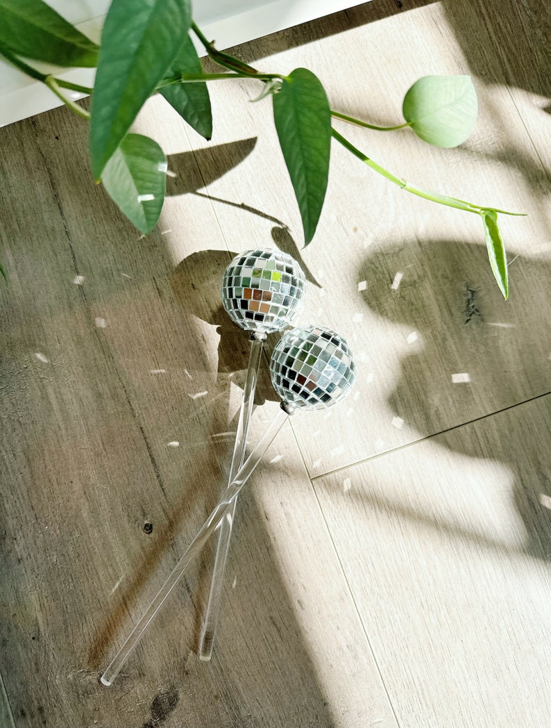 May include: Two silver disco ball ornaments on clear acrylic sticks. The sticks are crossed and the ornaments are facing each other. The ornaments are on a light brown wood floor.