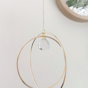 May include: A gold metal hoop mobile with a crystal prism hanging from the center. The mobile is suspended from a gold chain.