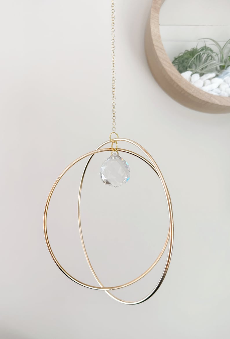 May include: A gold metal hoop mobile with a crystal prism hanging from the center. The mobile is suspended from a gold chain.