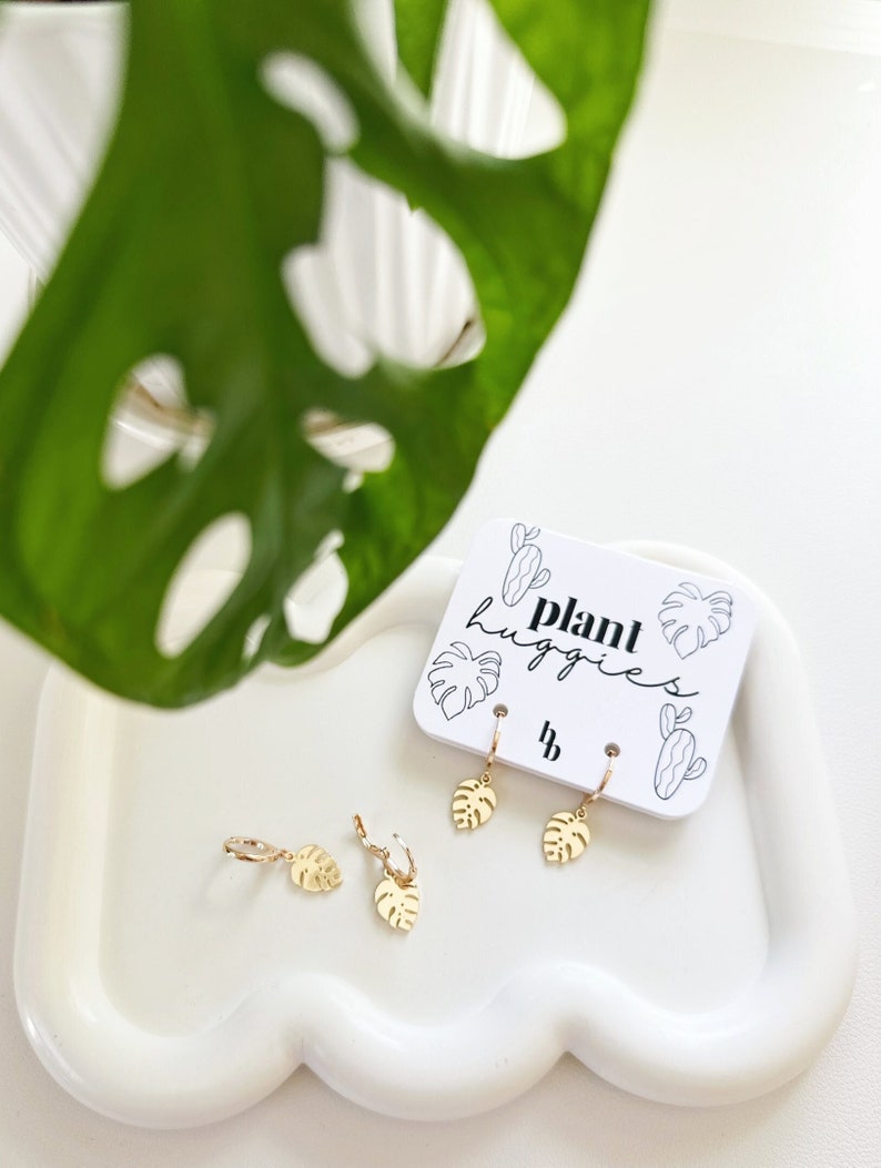 May include: Gold-toned earrings with a leaf design. The earrings are labeled "plant huggies".
