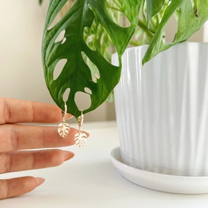 May include: Gold hoop earrings with a small, delicate monstera leaf charm hanging from each hoop.