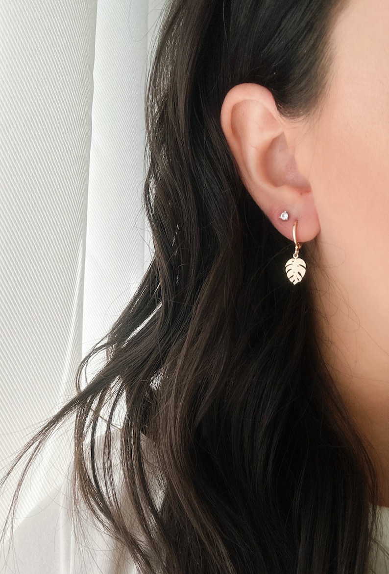 May include: A pair of gold hoop earrings with a small, clear gemstone stud in the upper lobe and a dangling gold leaf charm on the lower lobe.