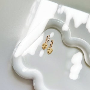 May include: A pair of gold hoop earrings with a small gold leaf charm hanging from each hoop. The earrings are on a white ceramic tray.