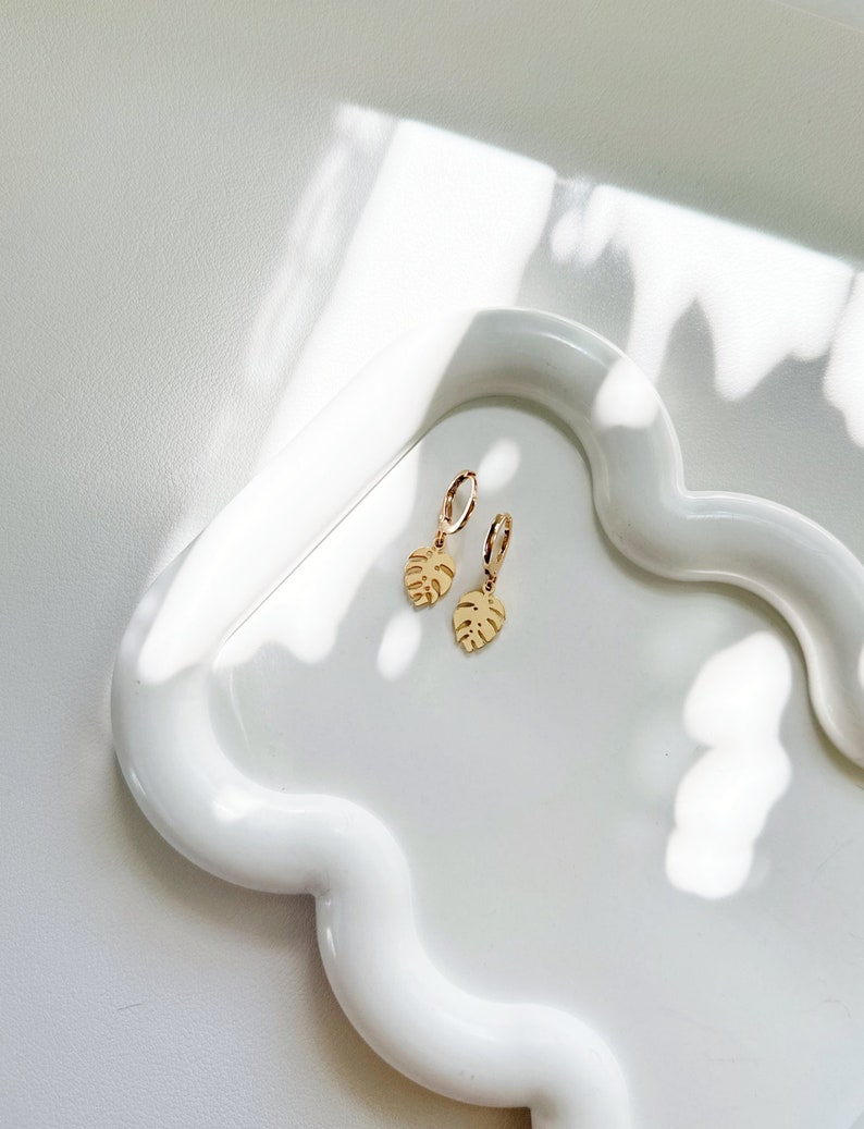 May include: A pair of gold hoop earrings with a small gold leaf charm hanging from each hoop. The earrings are on a white ceramic tray.
