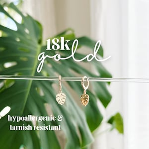 May include: A pair of gold hoop earrings with a small, delicate monstera leaf charm hanging from each hoop. The earrings are hypoallergenic and tarnish resistant. 18k gold.