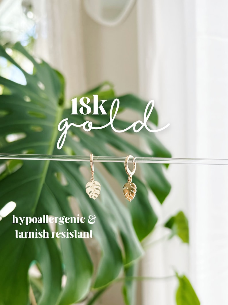 May include: A pair of gold hoop earrings with a small, delicate monstera leaf charm hanging from each hoop. The earrings are hypoallergenic and tarnish resistant. 18k gold.