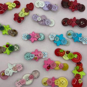 May include: A collection of hair clips decorated with assorted plastic buttons in various shapes, sizes, and colors including red, green, blue, pink, yellow, and purple. Many clips feature small embellishments like ladybugs, bees, and strawberries.