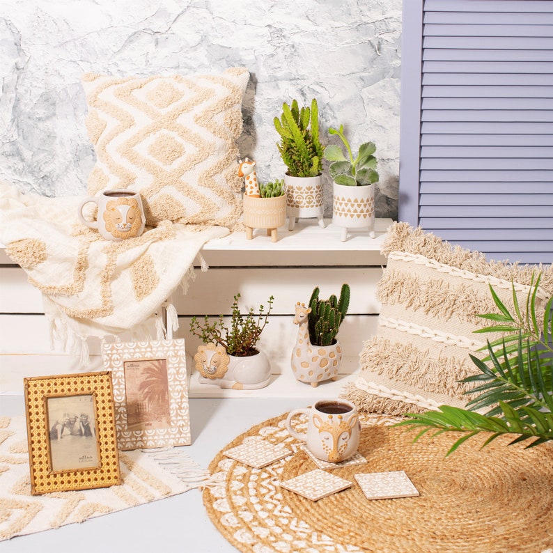 May include: A selection of safari-themed home decor. Features a beige patterned cushion, a coffee mug with a lion design, small planters with cacti and succulents, and a woven photo frame. The items are arranged on a white shelf and a jute placemat.