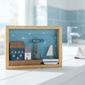 May include: A rectangular wooden shadow box frame containing a coastal scene with a blue background, white seagulls, a small blue sign reading BEACH, two miniature houses, and a white sailboat with a blue stripe on a wooden base.
