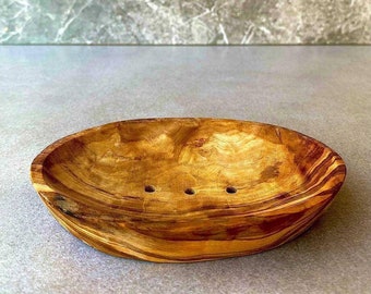 May include: Oval wooden soap dish with three drainage holes. The dish is made of light brown wood with darker brown grain patterns. The dish is set against a gray background with a marble-like texture.