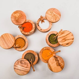May include: Wooden spice containers with lids and small scoops. The containers are filled with various spices in shades of orange, green, brown, and black. The containers are made of light brown wood with a natural grain.