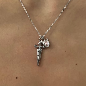 Peut inclure: Un collier en métal argenté doté d'un pendentif composé d'une petite breloque en forme d'épée et d'une breloque en forme de cœur avec une pierre précieuse rose clair. Le pendentif est suspendu à une fine chaîne argentée sur la clavicule.