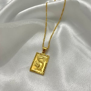 Puede incluir: Un collar de cadena dorada con un colgante rectangular que presenta un diseño de dragón detallado.