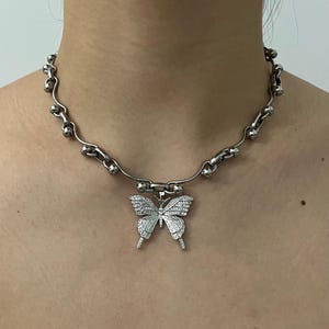 May include: Silver necklace with a butterfly pendant. The pendant is adorned with small, sparkling stones. The chain has a distinctive design, incorporating curved links and small, round beads. The necklace is designed to be worn around the neck.