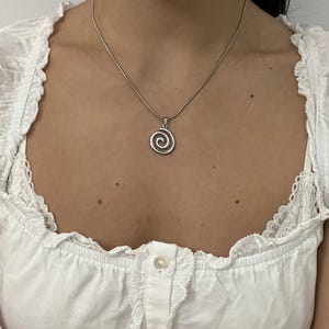 Pode incluir: Um colar de tom prateado com um pendente circular com um design de espiral em relevo. O pendente está suspenso numa corrente fina e lisa. O metal tem um acabamento polido que reflete a luz, criando uma aparência limpa para esta peça de joalharia.