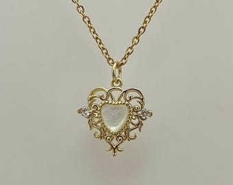 May include: A gold chain necklace with a heart-shaped pendant. The pendant features a white, iridescent stone surrounded by a delicate, filigree design. Two small, clear stones are set on either side of the stone.