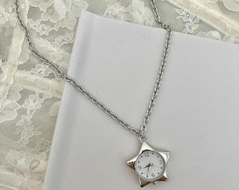 Op de afbeelding: Zilverkleurige ketting met een stervormige hanger met een horloge. De wijzerplaat van het horloge is wit met zwarte cijfers en wijzers. De ketting is fijn en de hanger is gecentreerd.