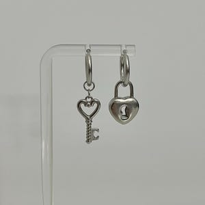 May include: A pair of silver-toned hoop earrings featuring mismatched charms. One earring has a dangling heart-shaped key charm, while the other earring features a heart-shaped padlock charm with a visible keyhole design on its front surface.