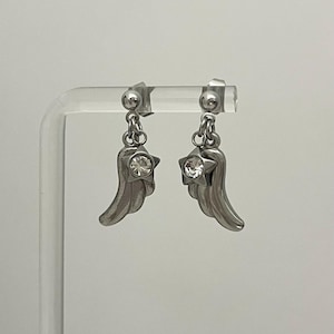 Op de afbeelding: Een paar zilverkleurige oorbellen met een kleine sterhanger met een heldere, ronde gefacetteerde kristal in het midden, bevestigd aan een gebogen vleugelvormige hanger. De oorbellen hebben een sluiting met een metalen bolletje.