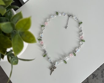 May include: A silver necklace with a fairy charm and clear, pink, and green beads. The necklace is on a white surface.