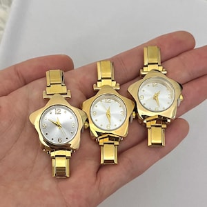 May include: Three gold-toned star-shaped watches with white faces and gold hands. The watches have a rectangular band and are held in a hand against a white background. The watch face displays the numbers 12, 3, 6, and 9.