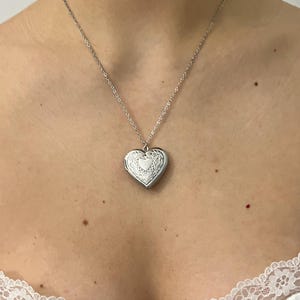 May include: A silver heart-shaped locket necklace. The locket features a detailed floral design around the edges and a smaller heart in the centre. The necklace chain is delicate and silver.