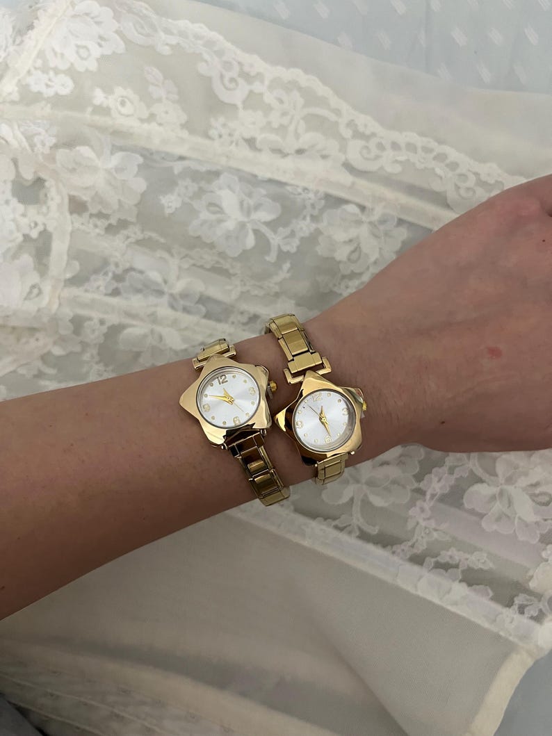 Pu&ograve; includere: Due orologi da polso color oro con quadranti a forma di stella e cinturini a maglie dorate. I quadranti degli orologi sono bianchi con lancette e indici dorati. Gli orologi sono indossati su un polso, su uno sfondo di pizzo bianco.