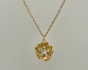 Puede incluir: Un collar dorado con un colgante de flor. El colgante tiene un grupo central de piedras preciosas transparentes y cinco pétalos. El collar cuelga de una fina cadena dorada. La imagen muestra el collar sobre un fondo liso.