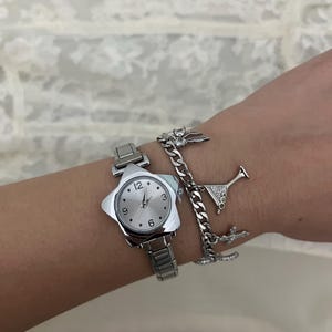 May include: A silver wristwatch with a star-shaped face and a matching silver strap. The watch face has silver hands and hour markers. A silver charm bracelet with a martini glass and other charms is also visible.