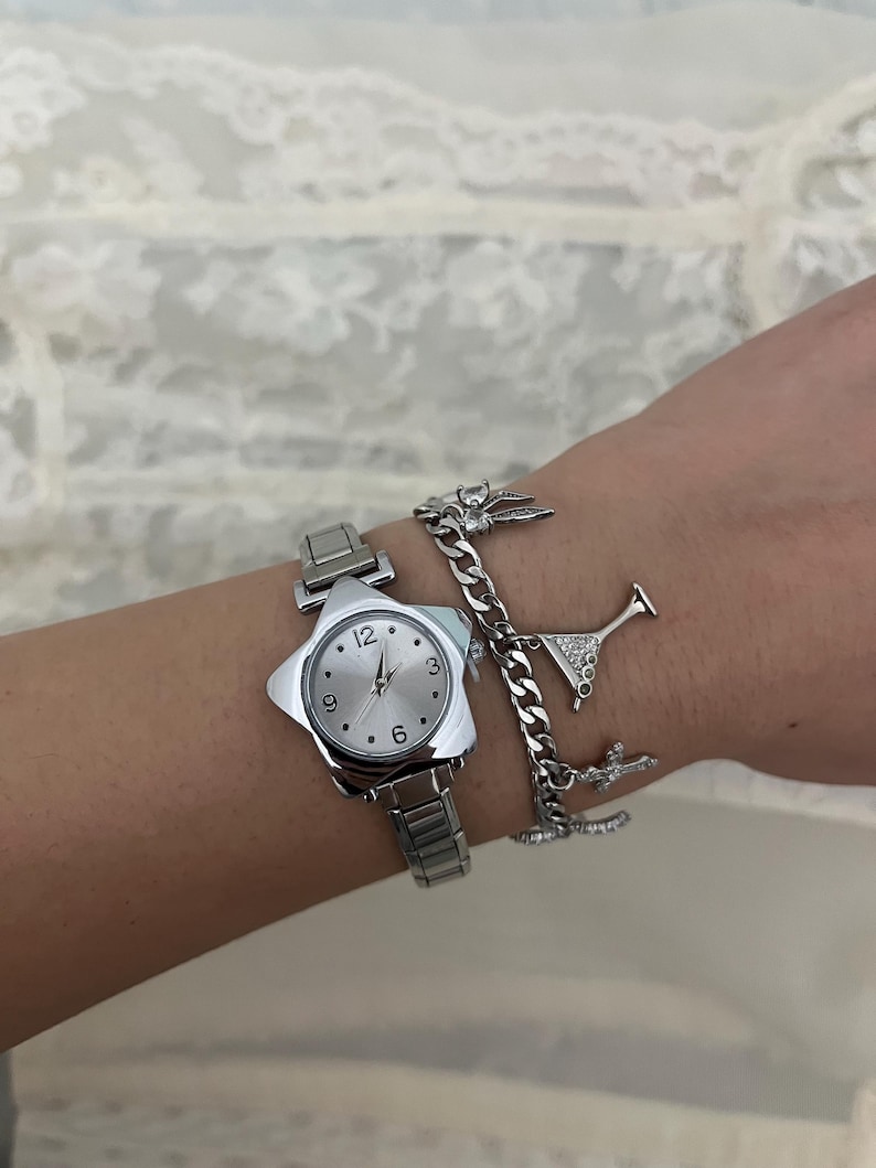 May include: A silver wristwatch with a star-shaped face and a matching silver strap. The watch face has silver hands and hour markers. A silver charm bracelet with a martini glass and other charms is also visible.
