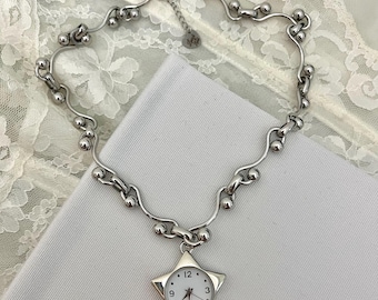 Op de afbeelding: Een zilverkleurige ketting met een stervormige horlogehanger. De ketting heeft een uniek ontwerp met kleine, ronde kralen en gebogen schakels. Het horloge heeft een witte wijzerplaat met zwarte cijfers en wijzers, en de tijd staat op 12:00 uur.